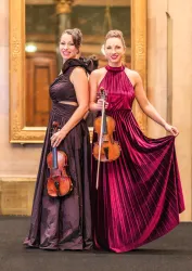 Zwei Musikerinnen stehen nebeneinander und halten Geigen in der Hand. Sie tragen elegante Kleider und lächeln in die Kamera.