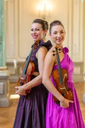 Zwei Musikerinnen stehen Rücken an Rücken und halten Geigen. Sie tragen elegante Kleider in Schwarz und Pink in einem schönen Raum.