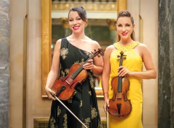 Zwei Musikerinnen stehen lächelnd in eleganter Kleidung. Sie halten Geigen und stehen vor einem geschmackvoll gestalteten Hintergrund.