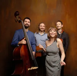 Eine vierköpfige Musikgruppe posiert vor einem braunen Hintergrund. Der Sängerin in gestreiftem Kleid stehen ein Kontrabass, eine Gitarre und eine Violine zur Seite.