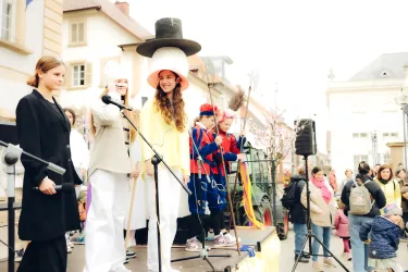 Eine Gruppe von Personen steht auf einer Bühne und hält Mikrofone. Sie tragen bunte Kostüme und sehen fröhlich aus, während eine Menge im Hintergrund zuschaut.
