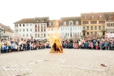 Ein großer Feuerhaufen brennt in der Mitte eines Platzes, umgeben von einer Menschenmenge. Die Zuschauer stehen gespannt und beobachten das Feuer.