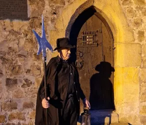 Ein Mann in schwarzer Kleidung steht vor einer alten Tür und hält einen langen Stock mit einer metallischen Spitze. Die Szene ist beleuchtet und vermittelt eine geheimnisvolle Atmosphäre.