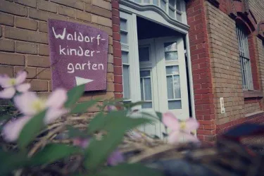 Ein Eingang zu einem Waldorfkindergarten mit einer türkisfarbenen Tür. Im Vordergrund sind einige Blumen sichtbar.