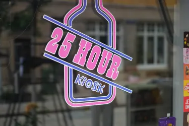 Ein bunter Aufkleber mit der Aufschrift "25 HOUR KIOSK". Der Hintergrund zeigt eine Stadtansicht mit Fenstern und Bäumen.