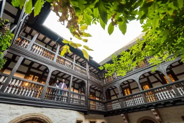 Ein historischer Innenhof mit Holzbalustraden und Pflanzen. Ein Mensch steht auf einem der Balkone und genießt die Atmosphäre.