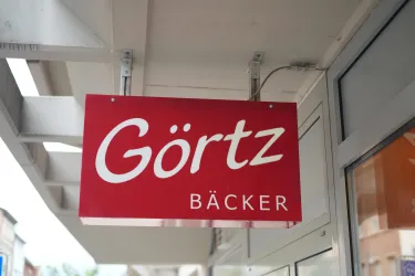Ein rotes Schild mit der Aufschrift "Görtz Bäcker". Es zeigt den Eingang einer Bäckerei.