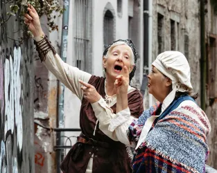 Zwei Frauen in traditionellen Kleidern stehen in einer engen Gasse. Eine Frau zeigt auf etwas und spricht begeistert, während die andere aufmerksam zuhört.