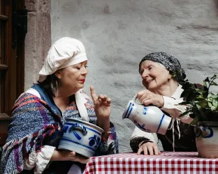 Zwei Frauen in traditioneller Kleidung unterhalten sich gemütlich an einem Tisch. Die eine hält einen Topf, während die andere mit einem Krug in der Hand zuschaut.