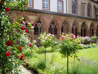 Ein schöner Garten mit bunten Rosen und einem gepflegten Rasen. Im Hintergrund sind historische Gebäudestrukturen mit kunstvollen Fenstern zu sehen.