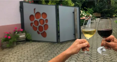Zwei Gläser Wein halten sich vor einem dekorativen Tor mit Traubenmotiv. Im Hintergrund sind Blumenkästen und eine grüne Umgebung zu sehen.