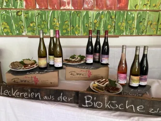 Eine Auswahl an Weinen steht auf Tischen, umgeben von verschiedenen Gerichten. Auf einer Tafel steht der Hinweis auf leckere Speisen aus der Bioküche.
