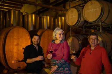 Drei Personen stehen in einem Weinkeller und halten Gläser mit Wein. Im Hintergrund sind große Holzfässer sichtbar.