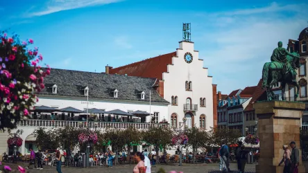 Ein belebter Platz mit einem historischen Gebäude im Hintergrund. Blumenampeln und viele Menschen verleihen der Szene eine lebendige Atmosphäre.