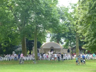 Ein Park mit vielen Bäumen und einer Menschenmenge, die vor einer Bühne sitzt. Es scheint eine Veranstaltung oder ein Konzert im Freien zu sein.