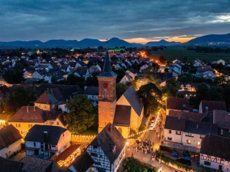 Eine malerische Stadtansicht bei Dämmerung, mit einer Kirche und beleuchteten Häusern. Im Hintergrund erheben sich sanfte Hügel.