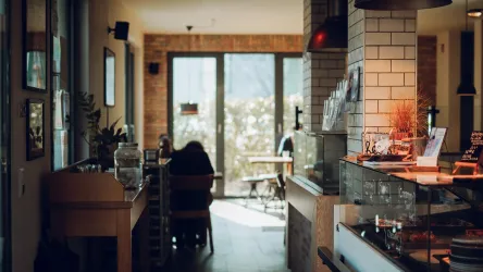 Ein modernes Café mit warmem Ambiente und gemütlichen Sitzplätzen. Große Fenster lassen viel Tageslicht herein und bieten einen Blick auf die Außenterrasse.