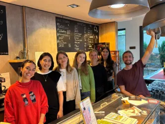 Eine Gruppe von fünf Frauen und einem Mann steht in einem Café hinter einer Theke. Auf der Wand sind Menütafeln sichtbar und die Atmosphäre wirkt freundlich und einladend.