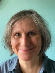 Eine Frau mit kurzen, grauen Haaren lächelt freundlich in die Kamera. Der Hintergrund ist in einem sanften Grünton gehalten.