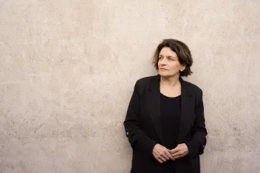 Eine Frau steht an einer grauen Wand und blickt nachdenklich in die Ferne. Sie trägt einen schwarzen Blazer und hat kurze, lockige Haare.