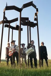 Eine Band steht im Freien vor einer modernen Skulptur. Die Bandmitglieder halten verschiedenen Musikinstrumente und tragen auffällige Outfits.