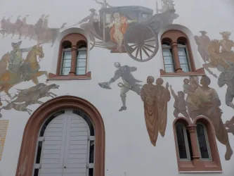 Eine Wand mit einem großen Wandgemälde, das historische Szenen zeigt. Es sind Figuren auf Pferden sowie Personen in verschiedenen Kleidungen dargestellt.