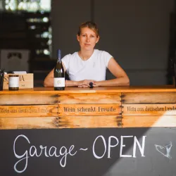Eine Frau sitzt an einem Holztresen mit Weinflaschen. Es steht "Garage - OPEN" an der Wand geschrieben.
