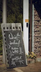 Ein Schild für den Weinverkauf steht vor einem Eingang. Es zeigt den Namen "Katrin WIND" und die Öffnungszeiten.