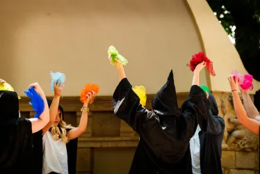 Eine Gruppe von Menschen trägt schwarze Gewänder und hebt bunte Tücher in die Luft. Sie scheinen an einer festlichen Veranstaltung oder Aufführung teilzunehmen.