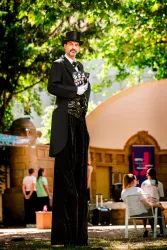 Ein maskierter Künstler steht auf Stelzen und trägt einen eleganten Anzug. Im Hintergrund sind Menschen und eine Baufassung zu sehen.