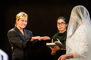 Eine Frau mit einer Krone und einem Anzug lächelt, während sie eine Geste macht. Eine andere Frau in einem Hochzeitskleid steht daneben, während eine dritte Person ein Buch hält.