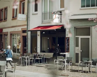 Ein gemütliches Straßencafé mit Tischen und Stühlen auf dem Bürgersteig. Im Hintergrund sind mehrere Gebäude mit modernen Fassaden zu sehen.