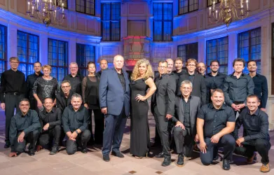 Eine große Gruppe von Menschen steht in einem eleganten Raum mit buntem Licht. Die Männer tragen schwarze Kleidung, während eine Frau in einem eleganten Kleid im Vordergrund posiert.