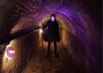 Eine düstere unterirdische Passage mit bunten Lichtern. Eine geheimnisvolle Figur steht am Ende des Ganges.