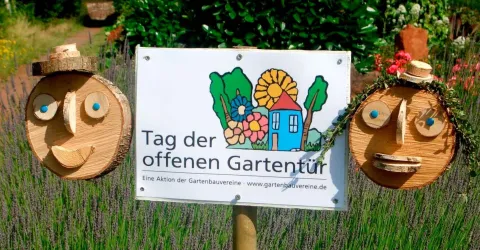 Ein Schild mit der Beschriftung "Tag der offenen Gartentür" steht zwischen zwei Holzgesichtern, die freundlich aussehen. Im Hintergrund sind bunte Blumen und Pflanzen zu sehen.