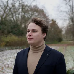 Ein junger Mann steht in einem winterlichen Park mit leicht schneebedecktem Boden. Er trägt einen eleganten Anzug und schaut nachdenklich in die Ferne.