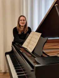 Eine Person sitzt an einem Klavier mit einem aufgeschlagenen Notenblatt. Im Hintergrund sind helle Vorhänge zu sehen.