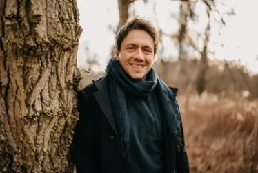 Ein lächelnder Mann steht an einem Baum in einer natürlichen Umgebung. Er trägt einen schwarzen Mantel und einen Schal.