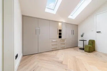 Ein modernes Zimmer mit einem großen, hellen Kleiderschrank und einem geflochtenen Parkettboden. Eine kleine grüne Sitzgelegenheit und ein Fenster sorgen für zusätzlichen Licht und Komfort.