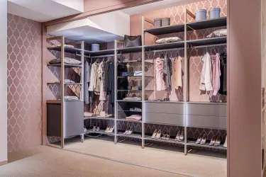 Ein moderner Kleiderschrank mit offenen Regalen und eine Vielzahl von Kleidungsteilen. Die Wände sind in einem zarten Rosa gehalten, was dem Raum eine elegante Ausstrahlung verleiht.