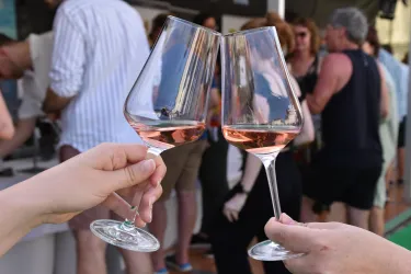 Zwei Weingläser mit Roséwein stoßen miteinander an. Im Hintergrund sind mehrere Menschen bei einer Veranstaltung zu sehen.