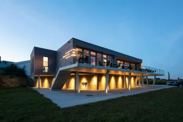 Ein modernes Haus mit großflächigen Fenstern und stilvoller Beleuchtung. Es steht auf Stelzen und ist von einer grünen Wiese umgeben.