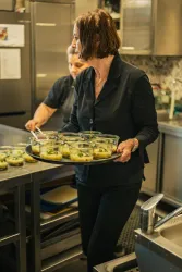Eine Frau trägt eine große Schüssel mit kleinen Portionen in einer modernen Küche. Im Hintergrund ist eine weitere Person mit Kochutensilien zu sehen.