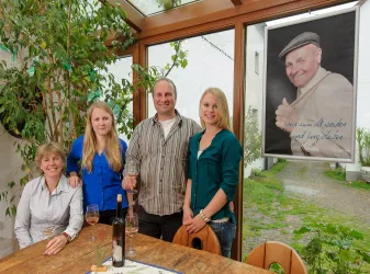 Eine Gruppe von vier Personen steht in einem hellen Raum mit Pflanzen im Hintergrund. Im Hintergrund hängt ein großes Porträt einer Person.
