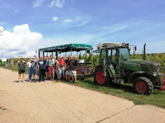 Eine Gruppe von Menschen steht neben einem Traktor und einem Weinfasswagen auf einem Feld. Der Himmel ist blau mit einigen Wolken.