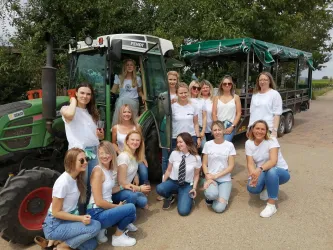 Eine Gruppe von Frauen in weißen T-Shirts posiert vor einem Traktor. Im Hintergrund ist eine grüne Planwagen zu sehen.