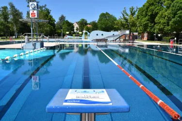 Ein Schwimmbad mit klarem Wasser und einem hohen Sprungturm. Im Hintergrund sind grüne Bäume und eine Rutsche sichtbar.