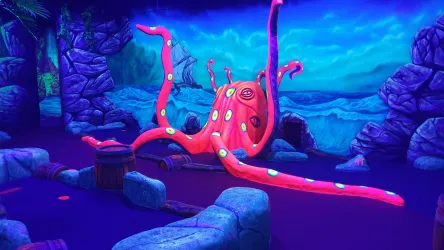 Ein großer, pinkfarbener Oktopus mit bunten Punkten in einer fantasievollen Unterwasserlandschaft. Die Umgebung ist mit Felsen und einem blauen, weiten Meer gestaltet.