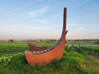 Eine große, stilisierte Schiffsskulptur steht inmitten eines Weinbergs. Im Hintergrund erstreckt sich der Himmel mit sanften Farben.