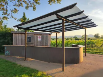 Ein moderner Unterstand mit einem abgedämpften Dach steht inmitten von Weinbergen. Im Hintergrund sind sanfte Hügel und Bäume zu sehen.